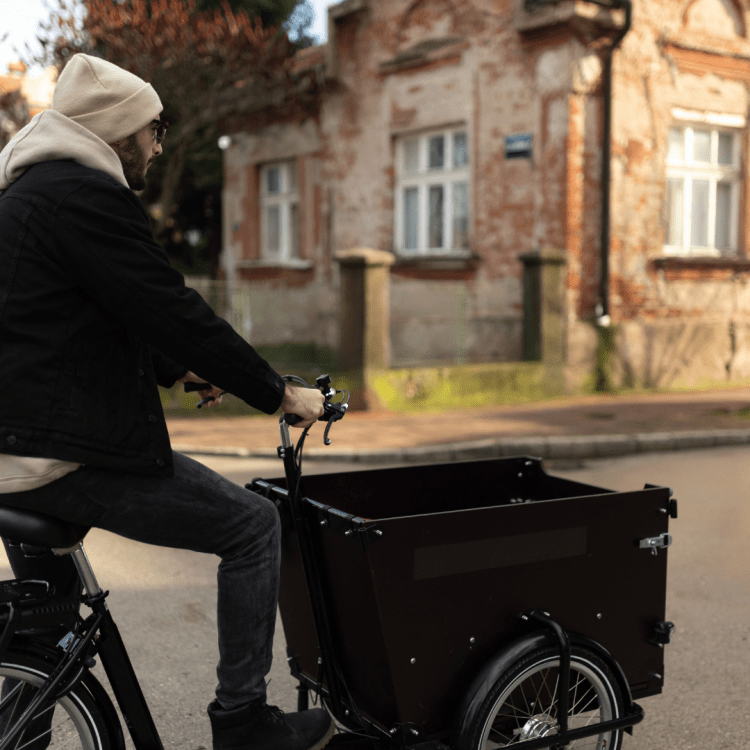 Man with glasses and wooly hat riding black cargo bike past a stone house.