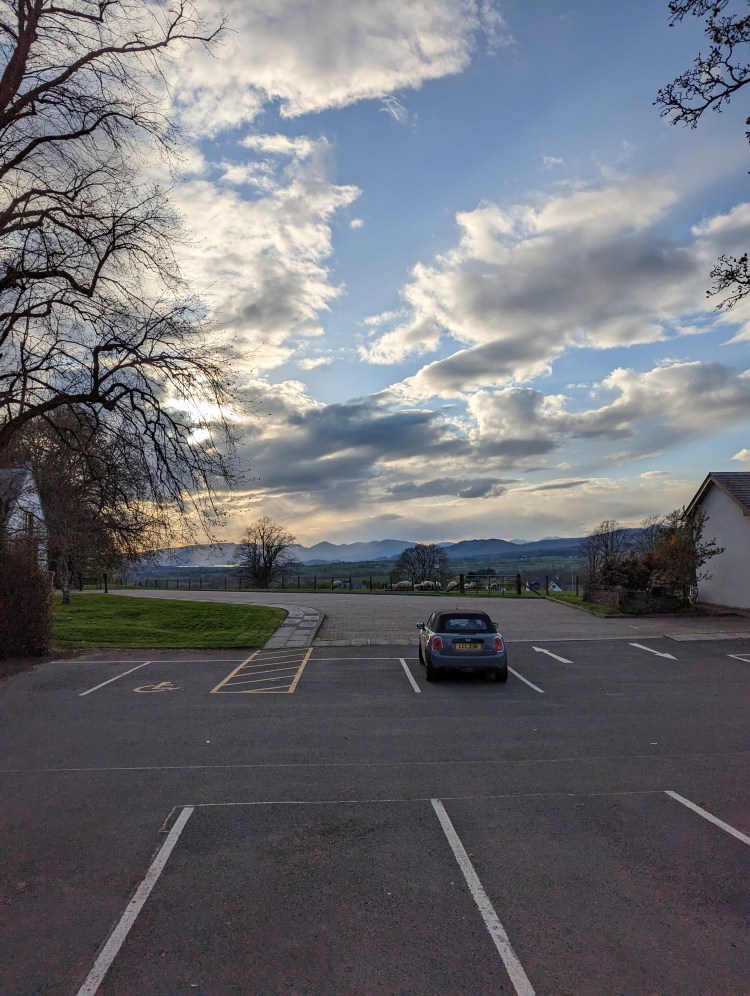 Car park with single soft top mini parked. Cloudy summer sky visible.