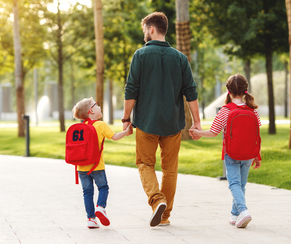 Man holding the hands of two young children as they walk along a wide path lined with trees